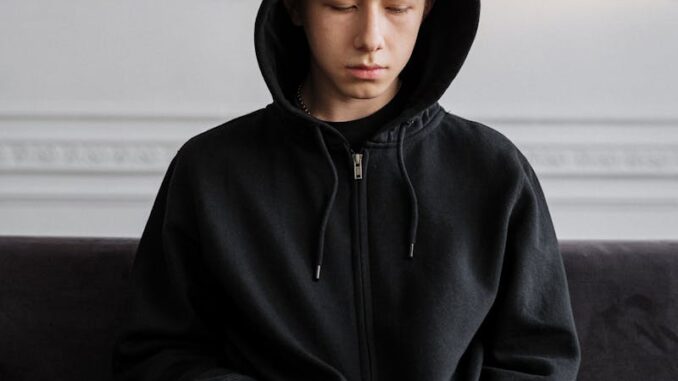 Teenager in a black hoodie sitting on a couch, reflecting on thoughts indoors.