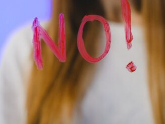 A teenager with long hair stands behind a glass with the word 'NO!' painted on it, against a blue background.