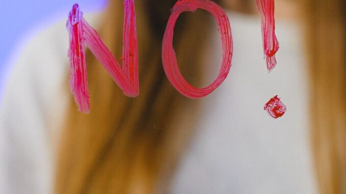 A teenager with long hair stands behind a glass with the word 'NO!' painted on it, against a blue background.