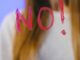 A teenager with long hair stands behind a glass with the word 'NO!' painted on it, against a blue background.